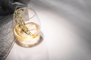 Şeffaf cam, bitki, buz küpü ve beyaz masada viski, kumaşın yanında da gölge var.
