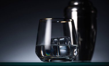 selective focus of transparent glass with ice cube and vodka in dark with back light and shaker clipart