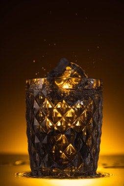 faceted glass with ice cube and splashing vodka in dark with warm back light clipart