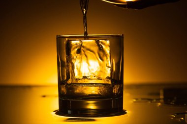 vodka pouring into transparent glass with ice cube in dark with warm back light clipart
