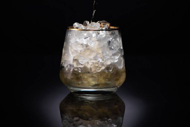 transparent glass with ice and pouring golden liquid on black background clipart