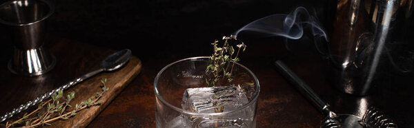 transparent glass with burning herb with smoke and ice cube on wooden surface, panoramic shot