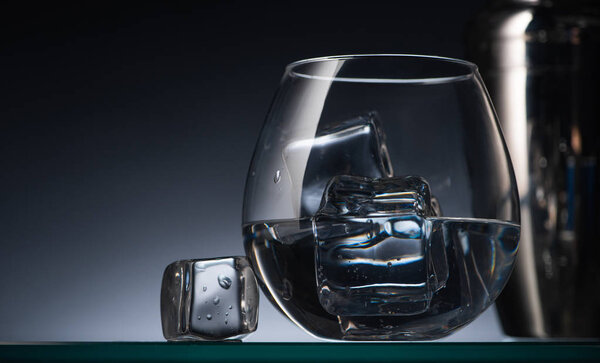 селективный фокус прозрачного стекла с кубиками льда и водкой в темноте с задним светом и шейкером
