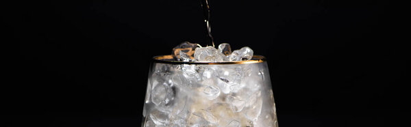 transparent glass with ice and pouring golden liquid isolated on black, panoramic shot