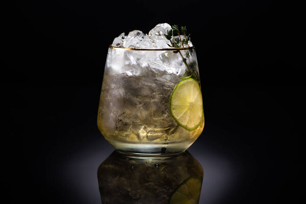 transparent glass with ice and golden liquid garnished with herb and lime on black background