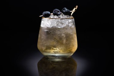 transparent glass with ice and golden liquid garnished with blueberries on black background clipart