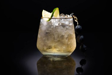 transparent glass with ice and golden liquid garnished with lime and blueberries on black background clipart