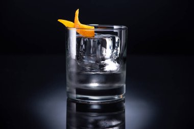 transparent glass with ice cube and vodka garnished with orange peel on black background clipart