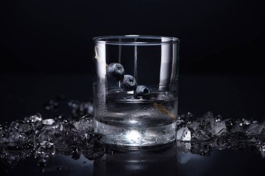 transparent glass with vodka and blueberries near smashed ice on black background clipart