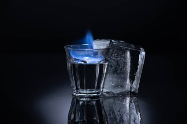 transparent glass with burning liquid near ice on black background clipart