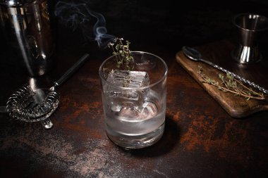 Yanan bitkilerle şeffaf cam, buz küpü ve yıpranmış yüzeyde votka.