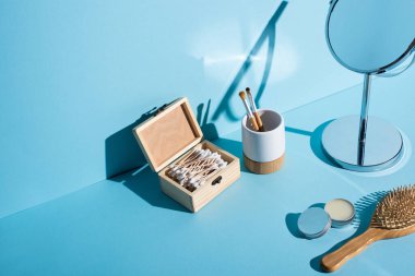 Toothbrush holder with cosmetic brushes, box of ear sticks, mirror, jar of wax and hair brush on blue background, zero waste concept clipart