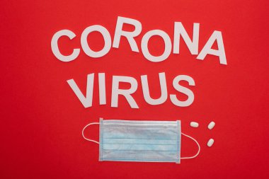 Koronavirüs harflerinin üst görüntüsü, tıbbi maske ve kırmızı üzerine izole edilmiş haplar.