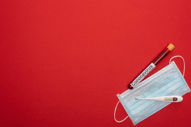 Kan örneği ve koronavirüs harfleriyle test tüpünün üst görüntüsü, kırmızı arka planda tıbbi maske ve termometre.