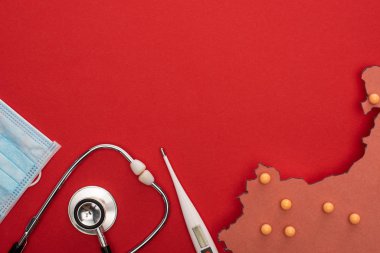 Termometrenin, steteskopun, tıbbi maskenin ve kırmızı zemin üzerinde sarı raptiyeler bulunan Çin haritasının üst görüntüsü