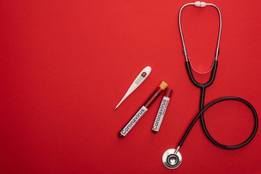 Stetoskop, termometre ve kan örnekleri ve kırmızı arkaplanda Coronavirus harfleriyle test tüplerinin üst görüntüsü