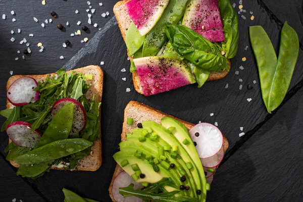 top view of fresh toasts with radish and avocado on stone board with pepper and salt