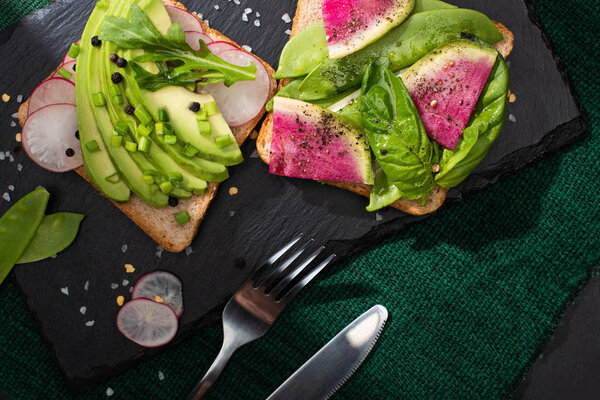 top view of vegetarian sandwiches with fresh vegetables on cloth and stone board with fork and knife