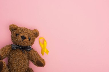Pembe arka planda oyuncak ayının yanındaki sarı kurdelenin üst görüntüsü, uluslararası çocukluk kanseri günü konsepti.