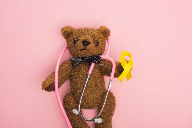 Top view of stethoscope with yellow ribbon on brown teddy bear on pink background, international childhood cancer day concept clipart
