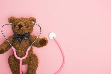 Pembe arka planda oyuncak ayıyla steteskopun üst görüntüsü, uluslararası çocukluk kanseri günü konsepti