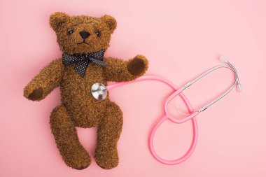 Pembe arka planda oyuncak ayıyla bağlantılı steteskopun üst görüntüsü, uluslararası çocukluk kanseri günü konsepti