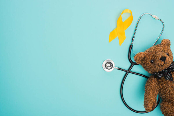 Top view of yellow ribbon, toy and stethoscope on blue background, international childhood cancer day concept