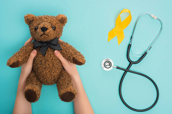 Cropped view of woman holding teddy bear near yellow awareness ribbon and stethoscope on blue background, international childhood cancer day concept
