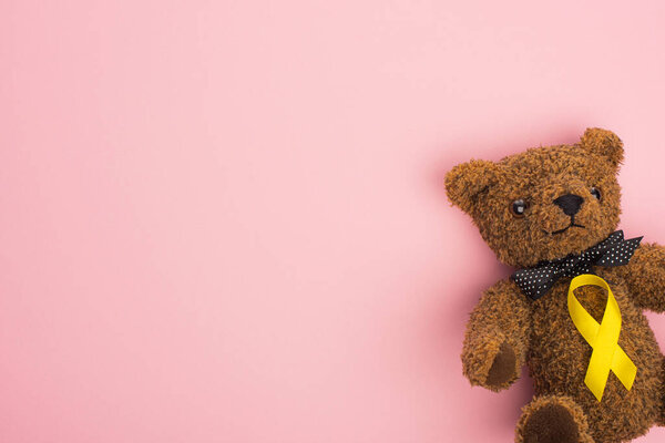 Top view of yellow ribbon on teddy bear with bow on pink background, international childhood cancer day concept