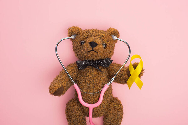 Top view of yellow ribbon and stethoscope on brown teddy bear on pink background, international childhood cancer day concept