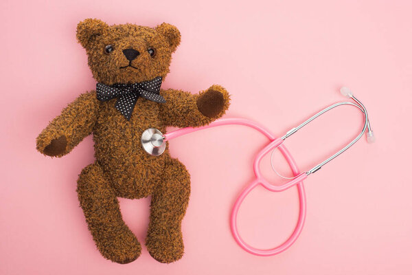 Top view of stethoscope connected with teddy bear on pink background, international childhood cancer day concept