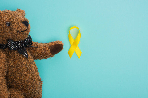 Top view of teddy bear with bow and yellow ribbon on blue background, international childhood cancer day concept