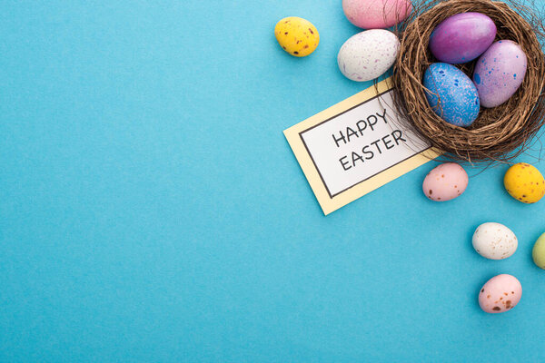 Top view of nest with painted chicken and perail eggs near card with happy easter lettering on blue background
