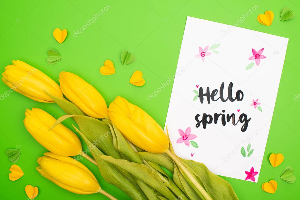 Top view of yellow tulips, card with hello spring lettering and decorative hearts on green background 