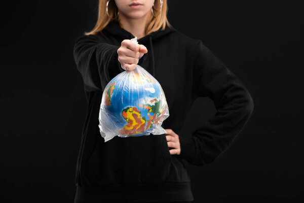 Partial view of woman with outstretched hand holding plastic bag with globe isolated on black, global warming concept