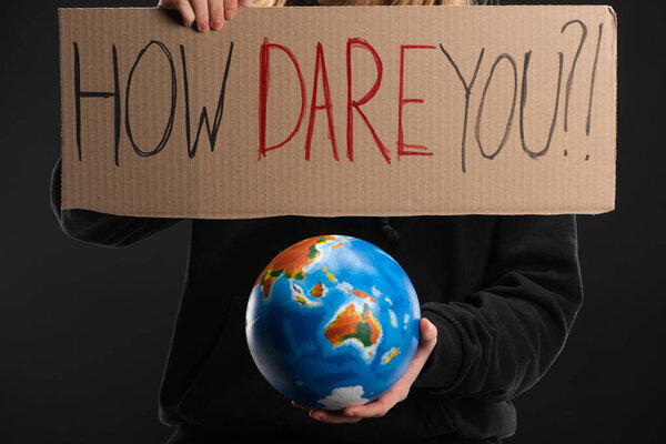 Partial view of woman holding globe and placard with how dare you lettering isolated on black, global warming concept