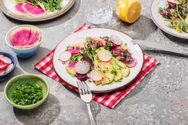 fresh radish salad with greens and avocado on napkin on grey concrete surface with ingredients in bowls and cutlery clipart