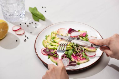 Yeşillikli taze turp salatası yiyen bir kadın ve beyaz yüzeyde su ile avokado yiyen bir kadın.