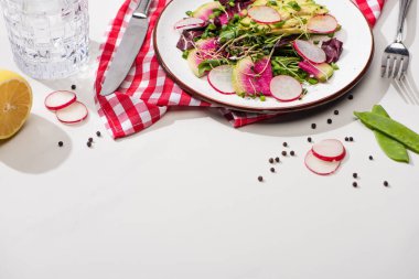 fresh radish salad with greens and avocado on plate on white surface with water and cutlery clipart
