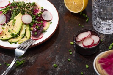 fresh radish salad with greens and avocado on plate on weathered surface with fork, lemon and water clipart