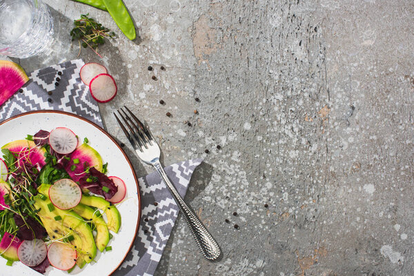 top view of fresh radish salad with greens and avocado on grey concrete surface with glass of water, fork and napkin