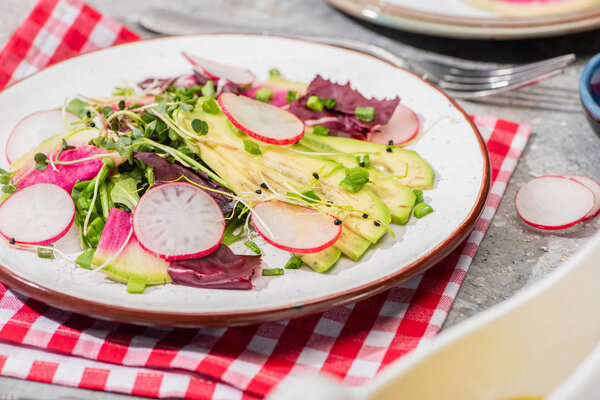 selective focus of fresh radish salad with greens and avocado served on napkin with cutlery on grey concrete surface