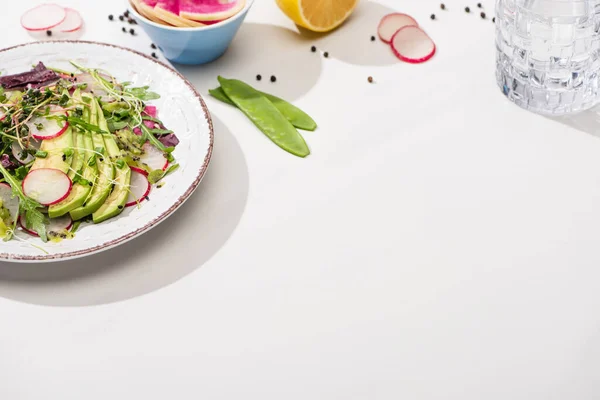 新鮮な大根のサラダと青菜とアボカドの白い表面に具材を入れたボウルと水 — ストック写真