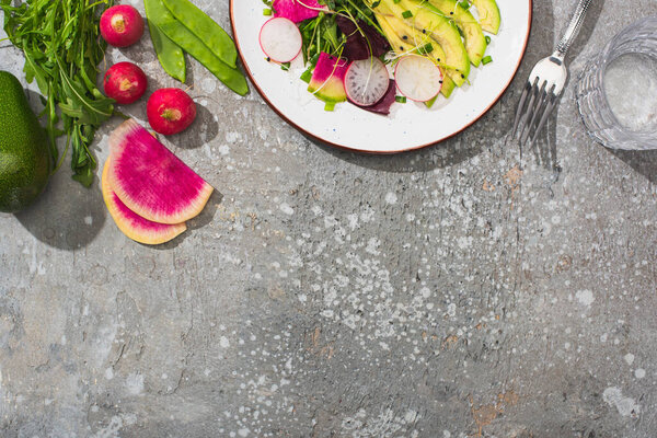 top view of fresh radish salad with greens and avocado near water, fork and vegetables on grey concrete surface