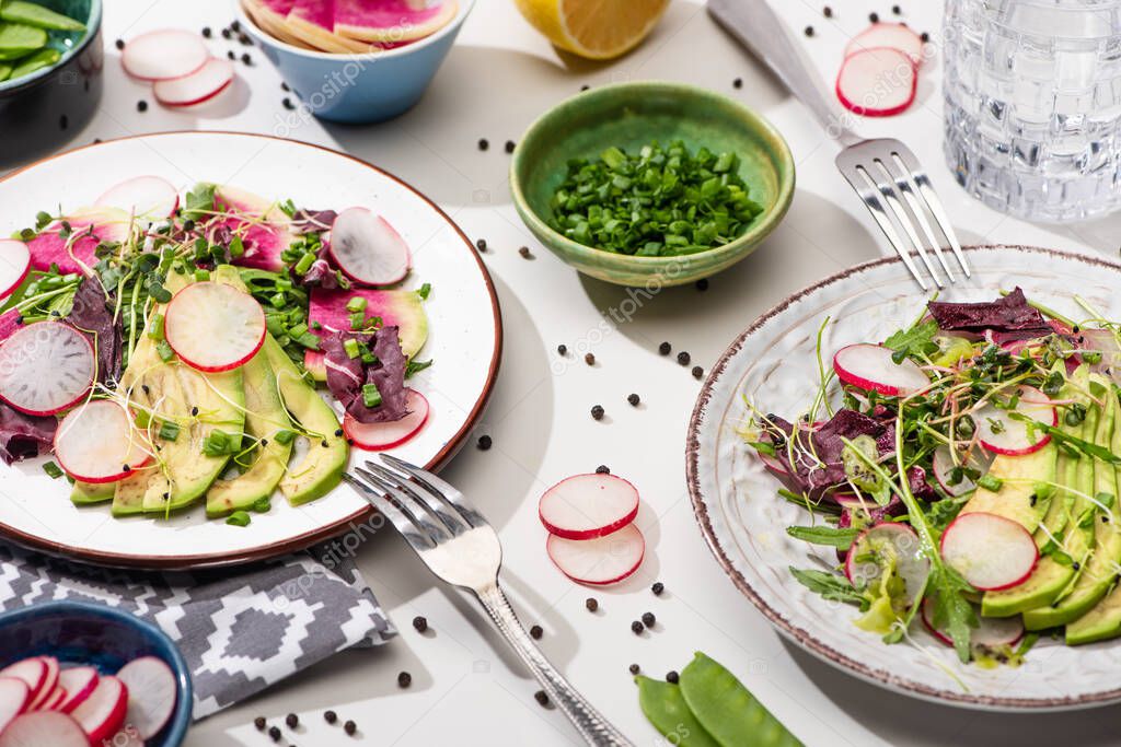 fresh radish salad with greens and avocado on plates on white surface with ingredients in bowls and water