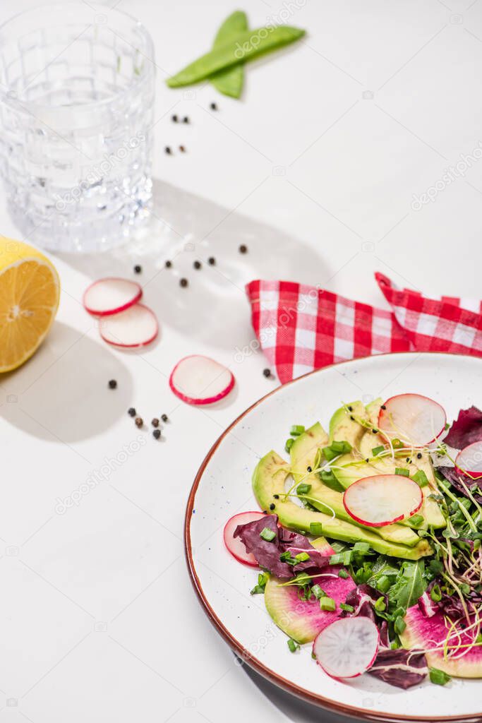 selective focus of fresh radish salad with greens and avocado on plate on white surface with water, lemon and napkin