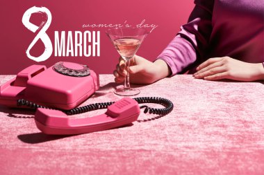 Kırpılmış görüntüde, 8 Mart 'ta izole edilmiş kadife kumaş üzerinde retro telefonun yanında bir bardak gül şarabı tutan kadın görülüyor.