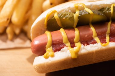 close up view of delicious hot dog with pickles, ketchup, mustard near french fries on wooden table clipart