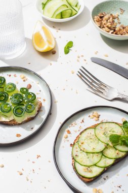 Tohumları, nane yaprakları ve limonu olan taze salatalıklı tostlar beyaz arka planda bir bardak su ve çatal bıçak takımı.