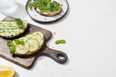 Taze salatalıklı tostlar ahşap kesim tahtasının üzerinde, suyun yanında ve limonlu beyaz arka planda